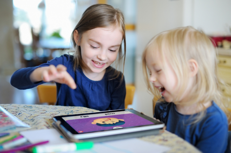 two girls laughing as they play a game together on a tablet