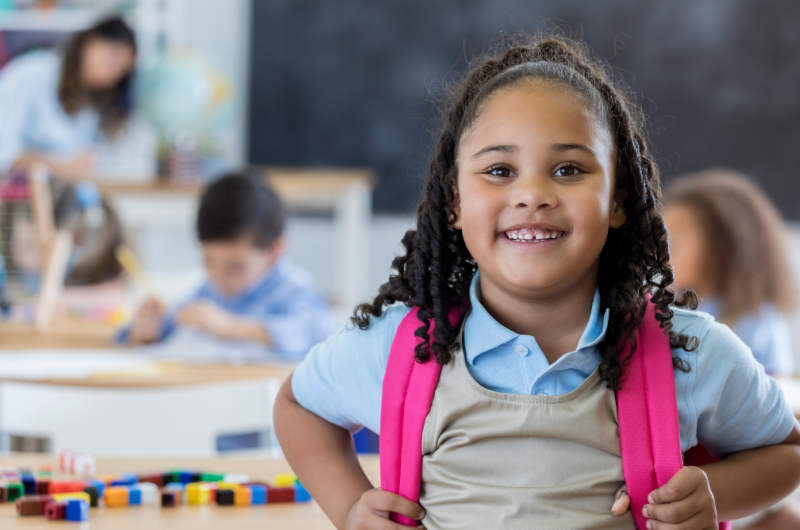 young black girl smiling with a backpack classroom in the background