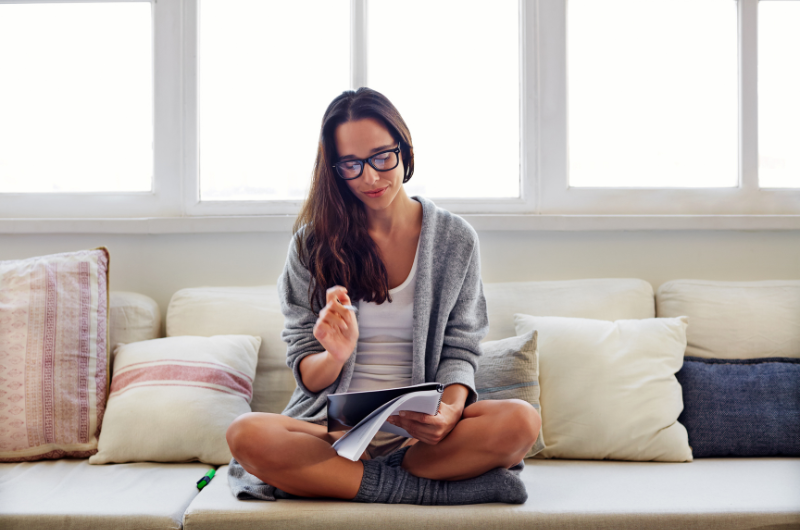 women sitting cross-legged on the couch in her lounge cloths reviewing a notepad