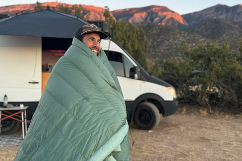 A man stands in the middle of a secluded wilderness area, wrapped comfortably in an Aeronaut Hoverquilt