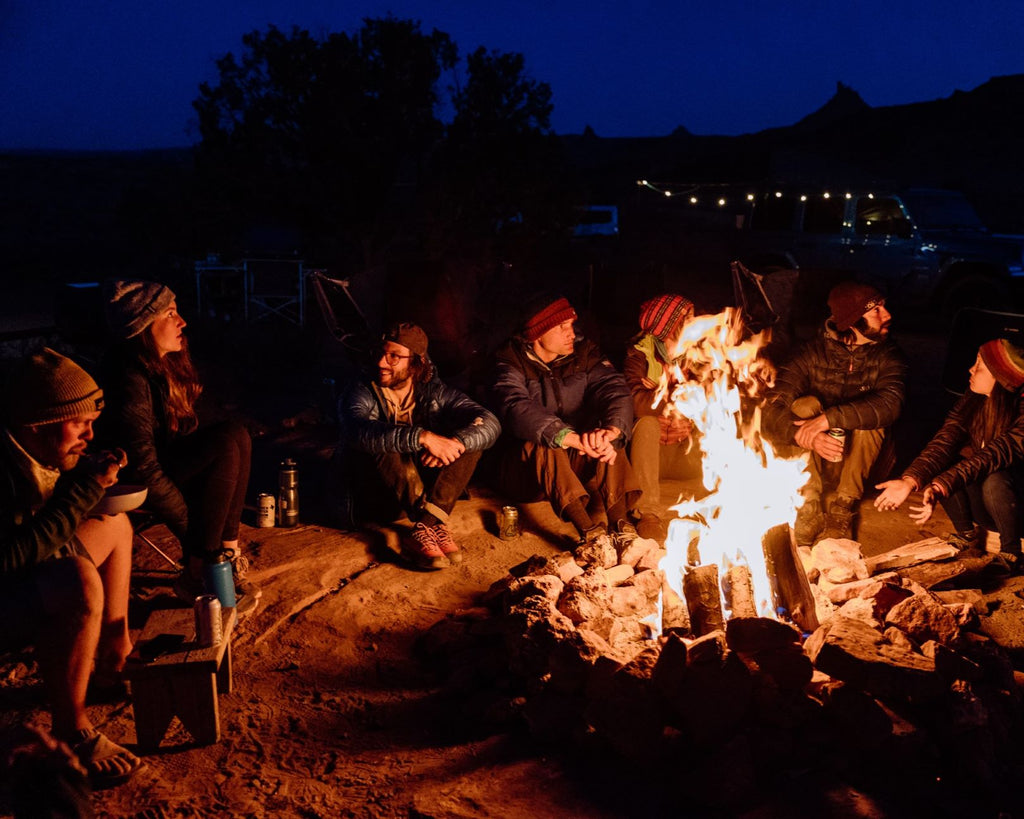 A group of people talk around a campfire at night. It's cold and they're wearing winter hats and jackets.