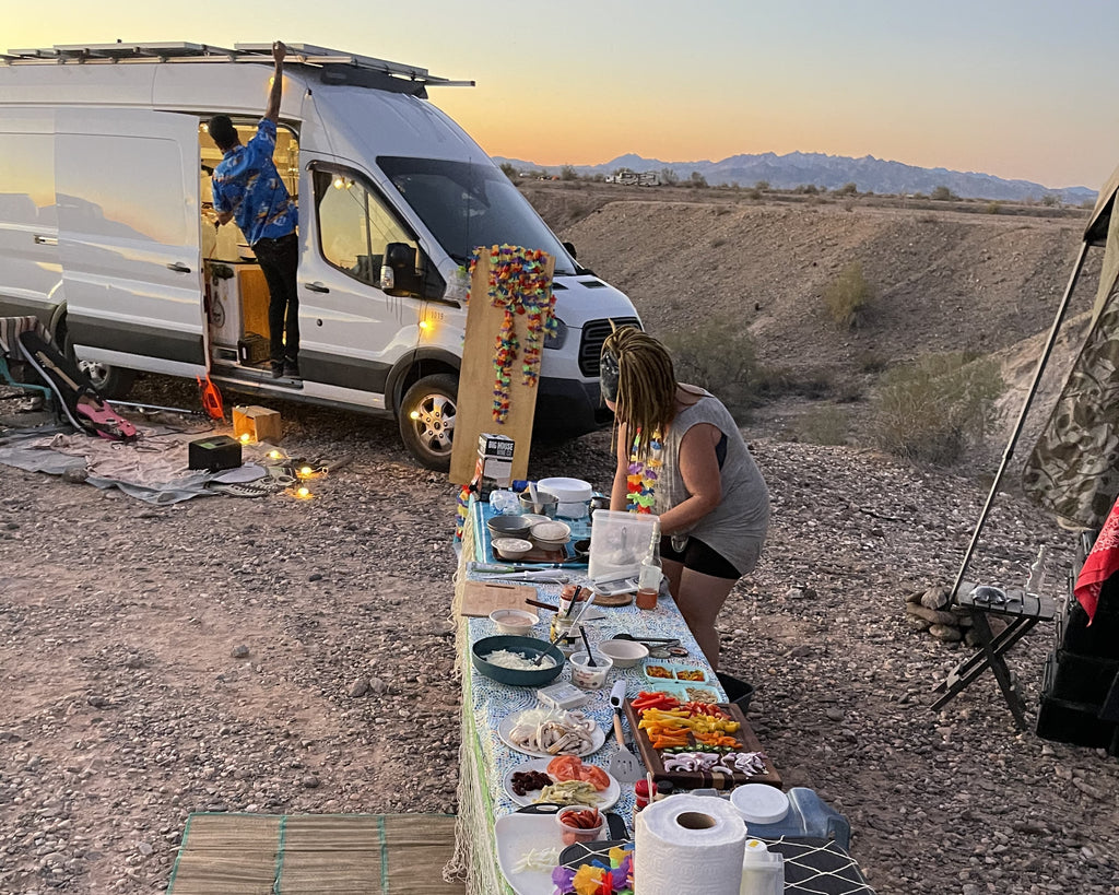 An outdoor camp scene. A woman prepares pizza toppings at a table while another person stands in the doorway of a Sprinter van.