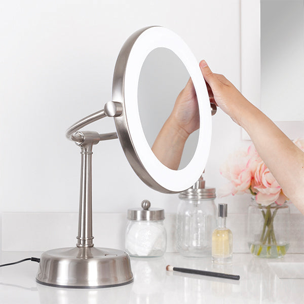 Zadro lighted makeup mirror with magnification with hand adjusting down the mirrorhead neck angle while on a white bathroom countertop next to sink and decorative jars