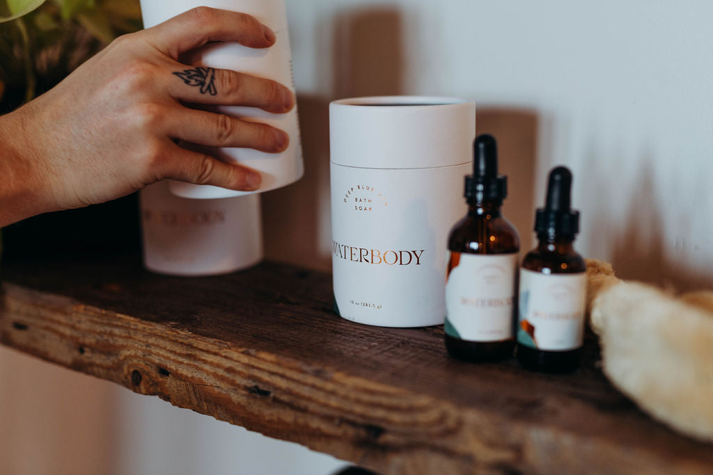 A hand places a tube of bath salts onto a bathroom shelf filled with Waterbody skincare products