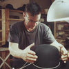 Mr. Pappa Hats, the Master Hatter of Agnoulita Hats, meticulously crafts a black hat by hand. In the background, wooden hat blocks and various tools are arranged on shelves, and a white desk lamp provides illumination.