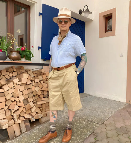 Tattooed gentleman sporting a classic fedora, striped shirt, and shorts, posing confidently with a European-style house backdrop.