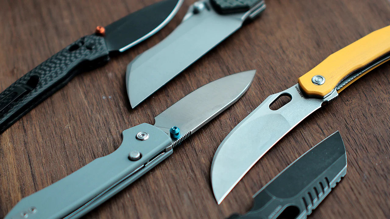 Collection of various folding knives with different blade shapes and handle designs arranged on a wooden surface, including models with yellow, black, and textured handles