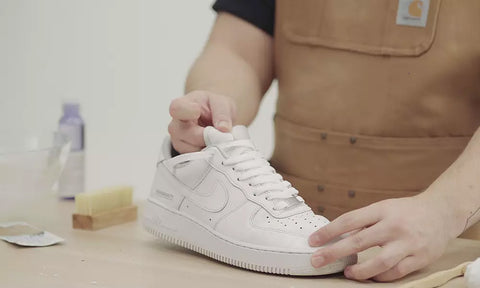 how to clean you sneakers featuring a man cleaning white sneakers on a light wooden table