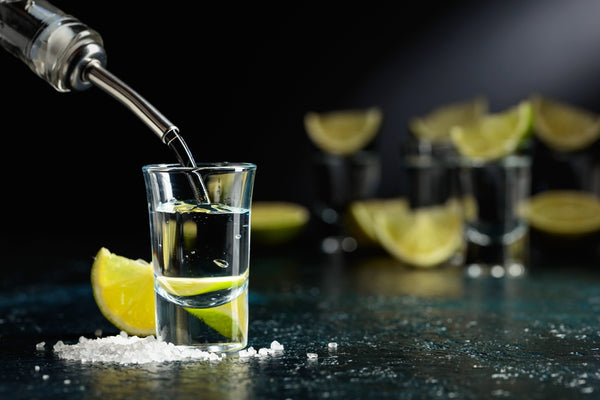 tequila being poured into shot glass with limes and salt and shot glasses in the background