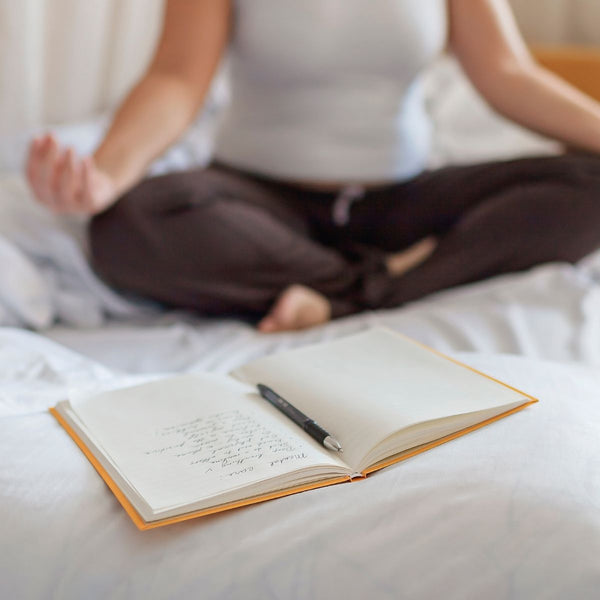 Tagebuch auf einem Bett, im Hintergrund meditiert eine Frau