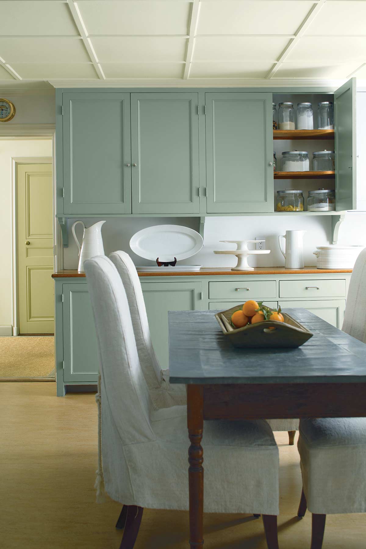 chairs around a kitchen table with fruit bowl centrepiece and light green cupboards as a backdrop