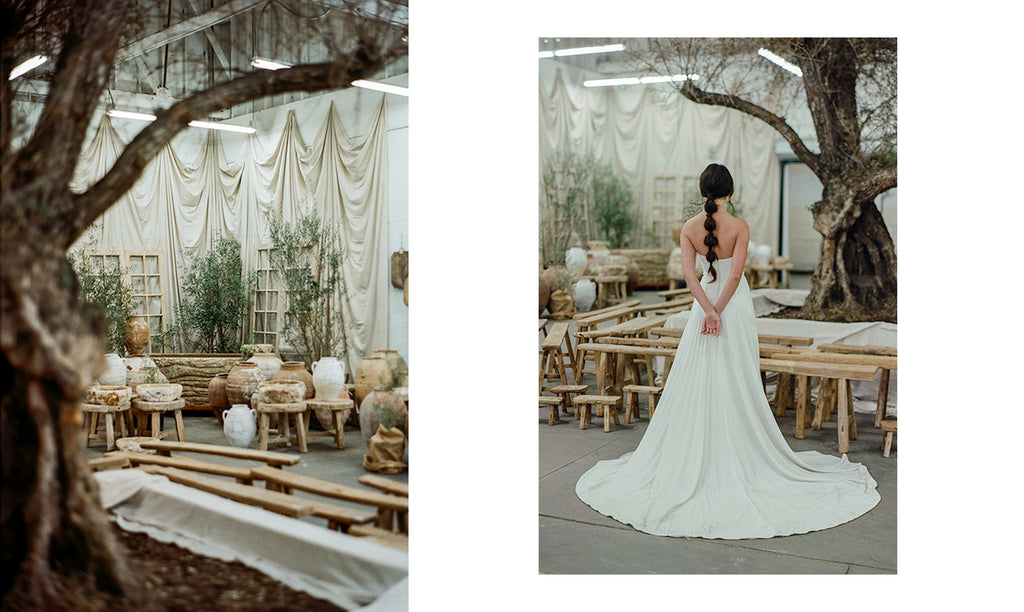 Model stands tall wearing a stunning white crepe wedding gown with long cathedral train facing an olive tree and ancient ceramic vessels