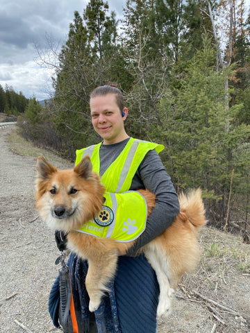 Julian is holding Runi; both are looking at the camera wearing neon yellow vests to pick up litter alongside the highway, which stretches away in the background with a border of evergreens.