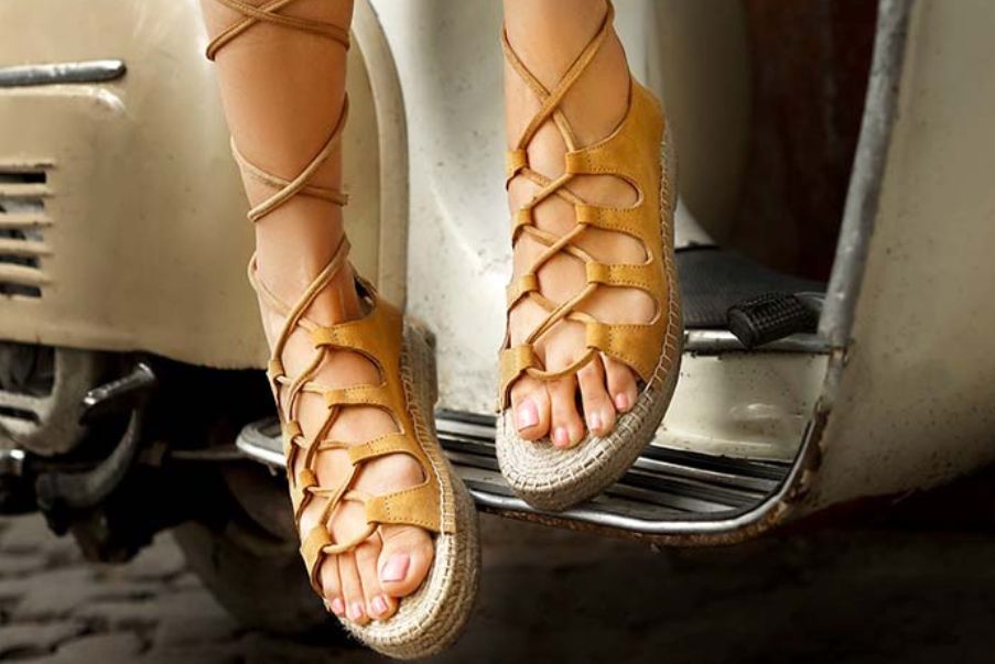 This is an image of a sandal for women which is beige in color wore by a girl model sitting on a scooter