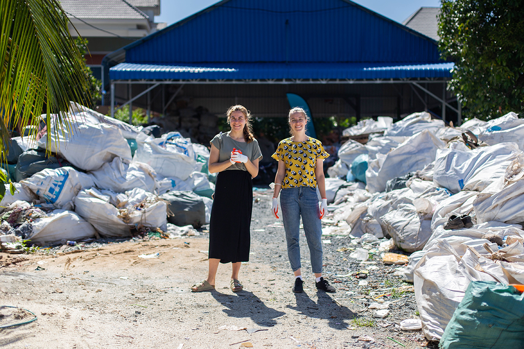 Katja und Maike stehen vor der Sortieranlage in Kambodscha. Um sie herum liegen viele Säcke Müll