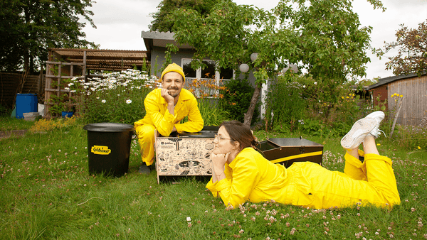 Zwei Personen in gelbem Overall liegen auf der Wiese im Garten, dazwischen sind Trockentoiletten von Goldeimer positioniert.