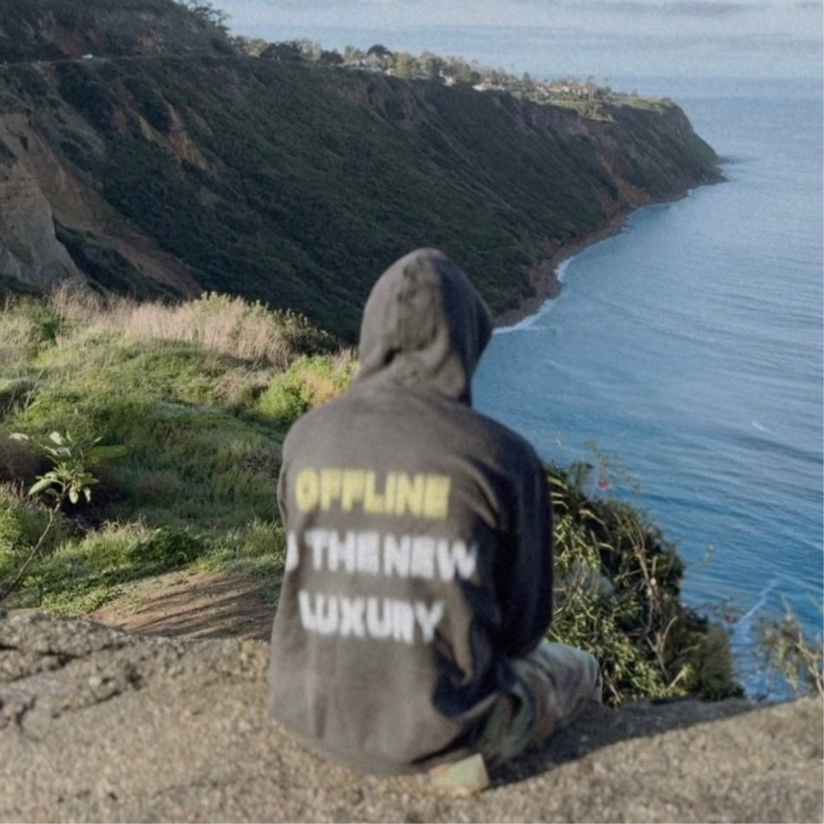 Person in hoodie overlooking a cliff and ocean, hoodie reads 'OFFLINE is the new luxury.'
