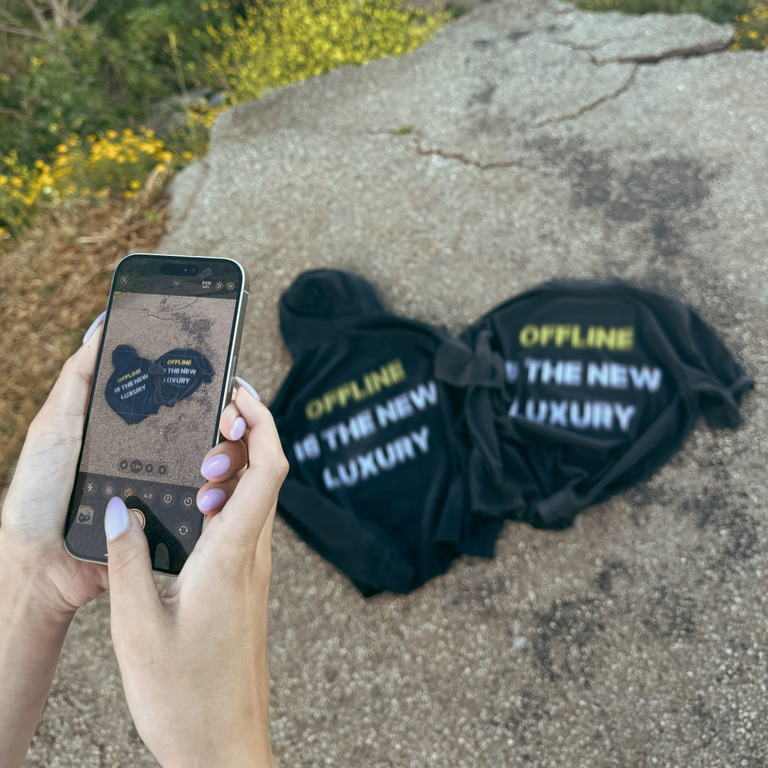 Hands holding a smartphone with a shirt onscreen matching the actual shirt on the ground, reading 'OFFLINE IS THE NEW LUXURY.'