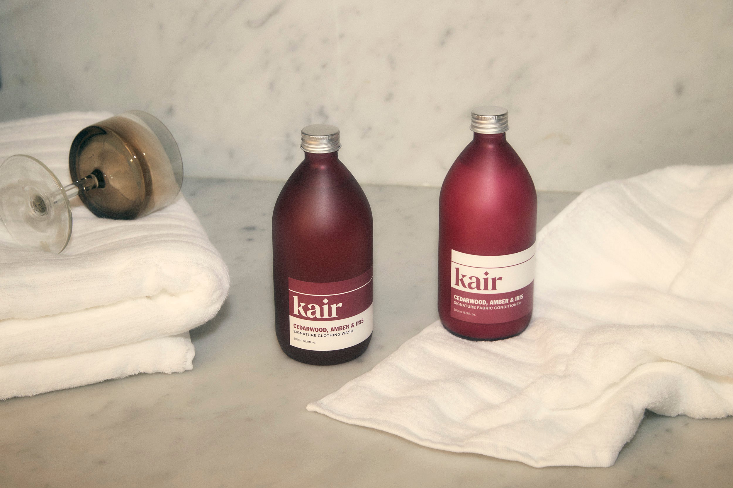 Kair Signature Fabric Conditioner - Cedarwood, Amber & Iris and Wild Juniper & Bergamot positioned among soft towels and a wine glass