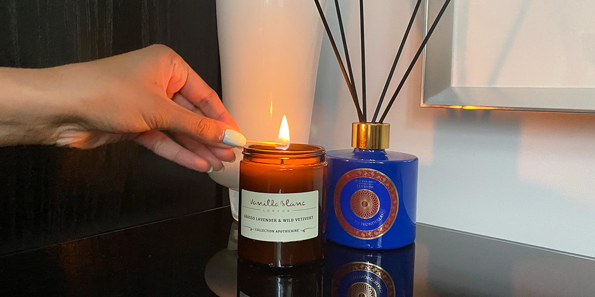 A hand holding a lit match over a candle on a glass table next to a diffuser and vase