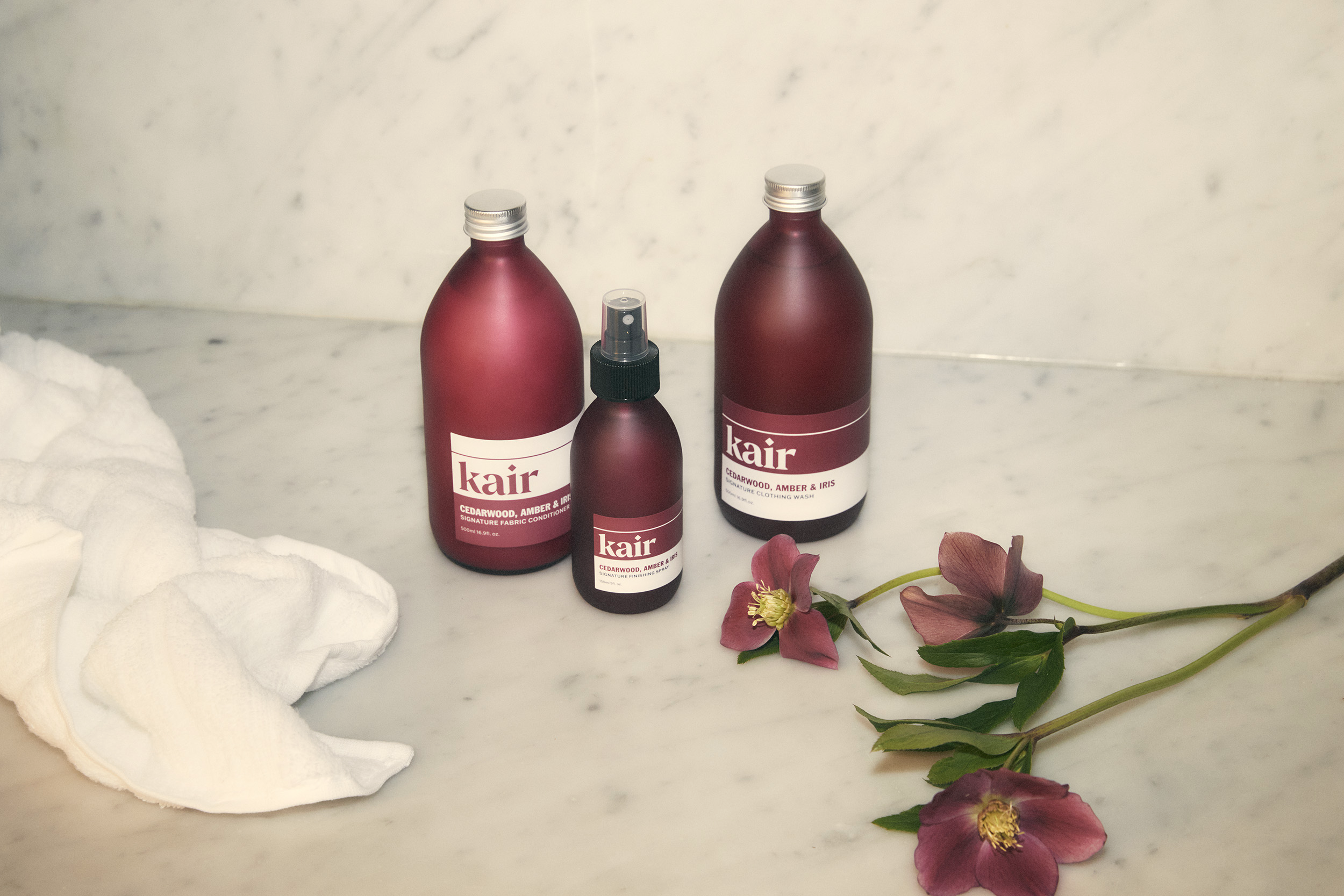 Photo showing Kair's Cedarwood, Amber & Iris Clothing Wash, Conditioner and spray on a white marble background next to some white towels and a flower