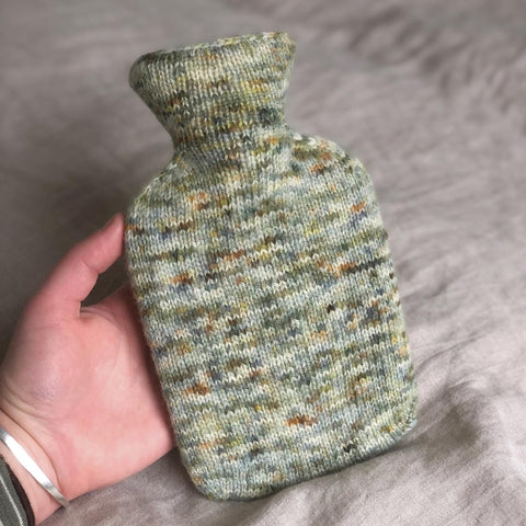 Small, multi coloured hot water bottle being held up by a white woman's hand on a linen sheet.