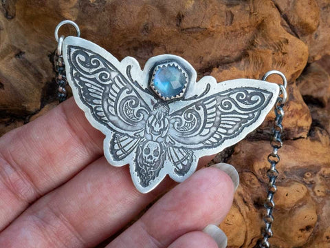 A sterling silver death head moth necklace with rainbow moonstone. The death head moth is etched into silver and cut out in a moth shape. It is suspended from the wing tips by a sterling silver rolo chain. A rainbow moonstone hex shaped stone is on top