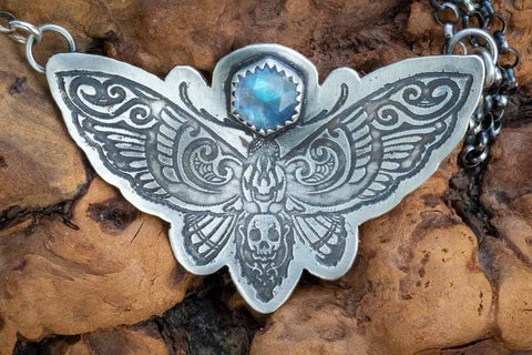 A death head moth necklace in sterling silver. The witchy style design is etched into the silver. A hex-shaped rainbow moonstone is set above the death head moth. The necklace is suspended from either wing tip with a sterling silver rolo chain.