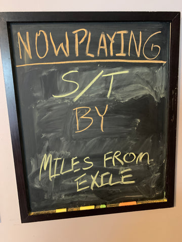 chalkboard with writing saying "now playing: s/t by miles from exile"