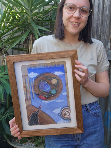 A short hair brunette, short to mid-height woman, wearing glasses. Holding a framed painting of a zoomed/cropped table with cutlery,  a plate with the universe on it and rocket
