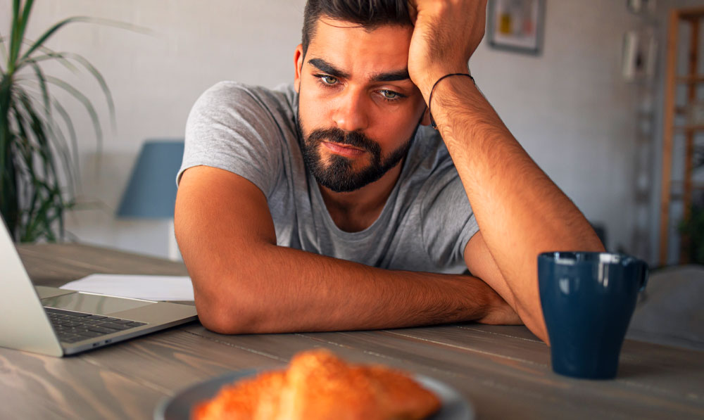 Frustrated man staring at breakfast with a poor appetite with laptop and coffee nearby