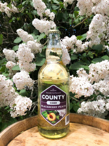 A bottle of blackberry peach county cider sitting on a barrel with flowers in the back