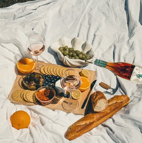 A photo of a picnic blanket with a charcuterie board of various foods and a bottle of wine on it