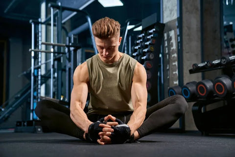 Man in black leggings and a green muscle shirt sitting on the floor of a gym in a butterfly position.