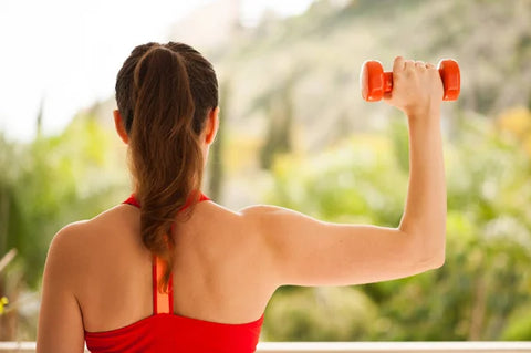 A woman in a red sports top holds a small red dumbbell in her hand as she holds her arm at shoulder height with her elbow bent so her forearm is pointing straight up and her hand is level with the top of her head with a tree covered hill in the background.