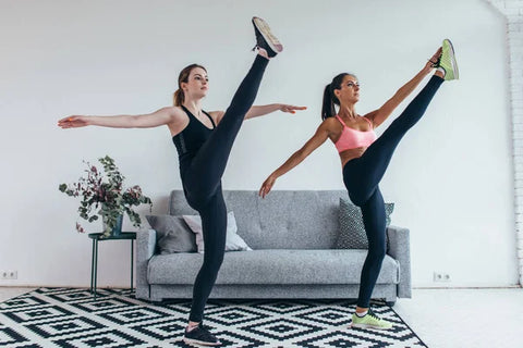 Two women wearing workout attire standing in a living room with one leg stretched up in a high kick. 