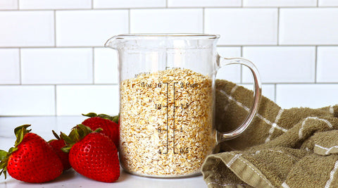 A measuring cup filled with oats, some strawberries and a towel with a white subway tile backdrop.