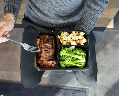 Person in a grey sweatshirt sitting holding a boxed meal of steak, potatoes, and broccoli and a fork.
