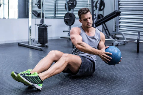 Man in a grey tank top and shorts sitting on the gym floor holding a medicine ball to one side.