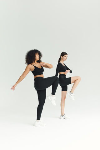 Two women in black workout attire each with one leg up with the thigh horizontal to the ground and the knee bent.