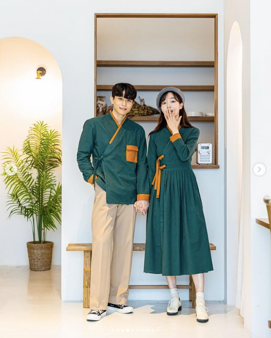 A couple wearing modern green hanboks in a white minimalist room with potted fern