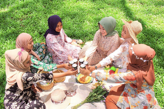 Six girls wearing hijabs and flowery dresses clinking glasses at a picnic