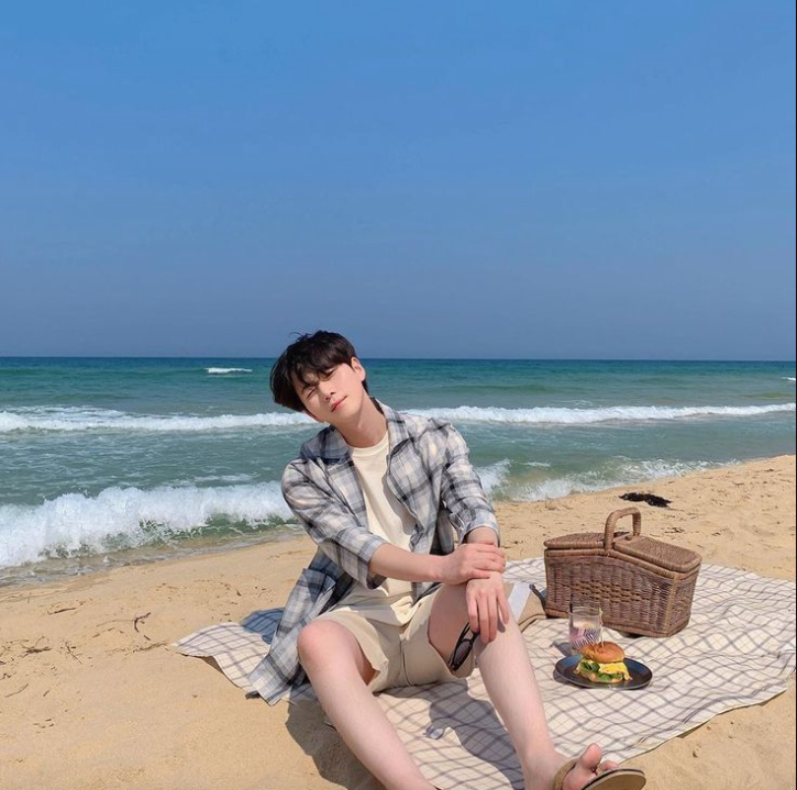 A man wearing a plaid unbuttoned button-up shirt by the ocean with a picnic basket