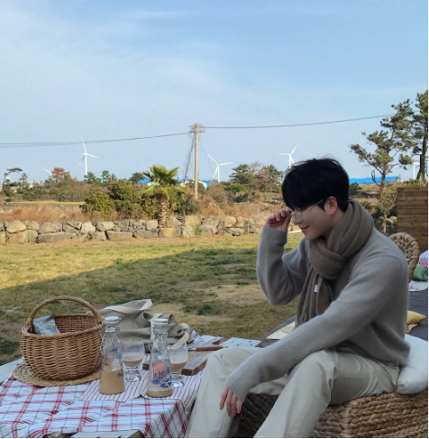 A man wearing a gray sweater with a scarf and cream trousers at a picnic site