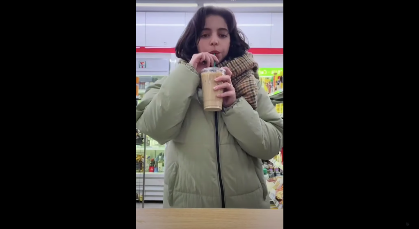 A girl drinking iced coffee in a beige coat in a convenience store