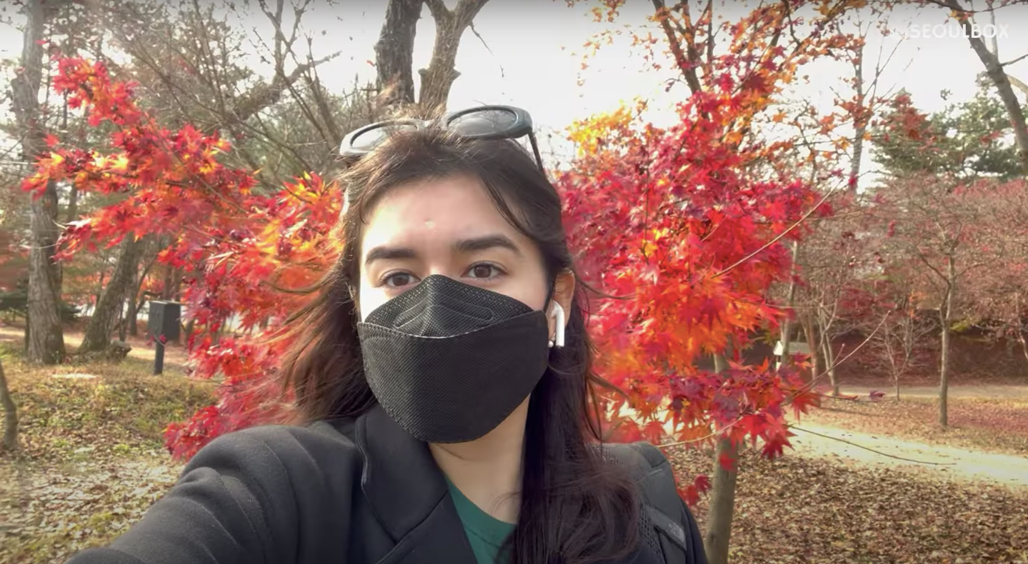 A woman wearing a mask in front of trees with red-orange leaves