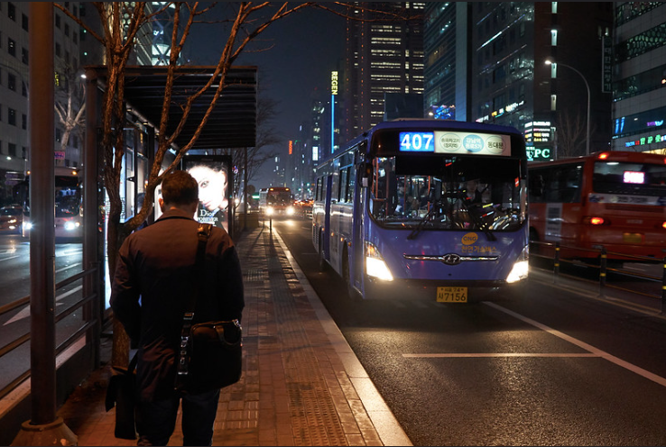 A man waiting at a bus stop for a blue city bus at dusk