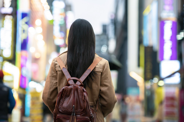 Back of woman wearing camel coat and pink backpack looking at neon-lit towers