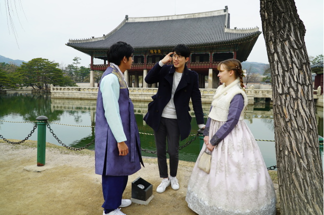 A man in a black blazer and modern clothes between two people in purple hanboks in front of palace