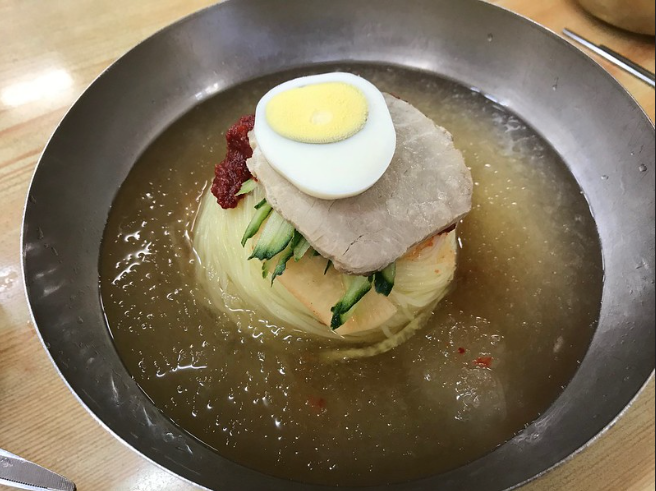 A metal bowl with a boiled egg, meat, and noodles in cold broth